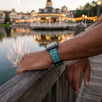 Teal icon-pattern watchband beside an illuminated waterfront pavilion